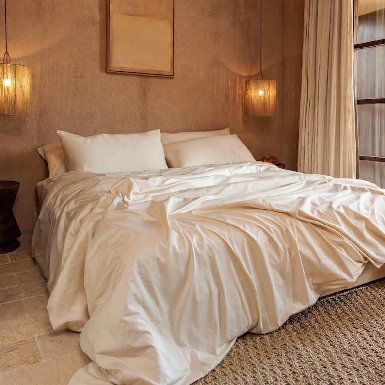 May include: A bed with cream-colored bedding, including a duvet cover and pillows. The bed is in a room with a neutral color palette, with a woven rug and a window with sheer curtains. Two pendant lights hang above the bed.