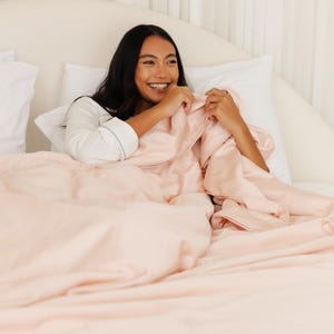 May include: A person in a white shirt with light blue trim is smiling while holding a light pink satin comforter and sheets. The bedding is on a white bed with white pillows.