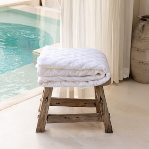 May include: A stack of folded white quilted blankets rests on a rustic wooden stool. The blankets have a diamond pattern. The stool is made of weathered wood. The background includes a swimming pool and a sheer curtain.