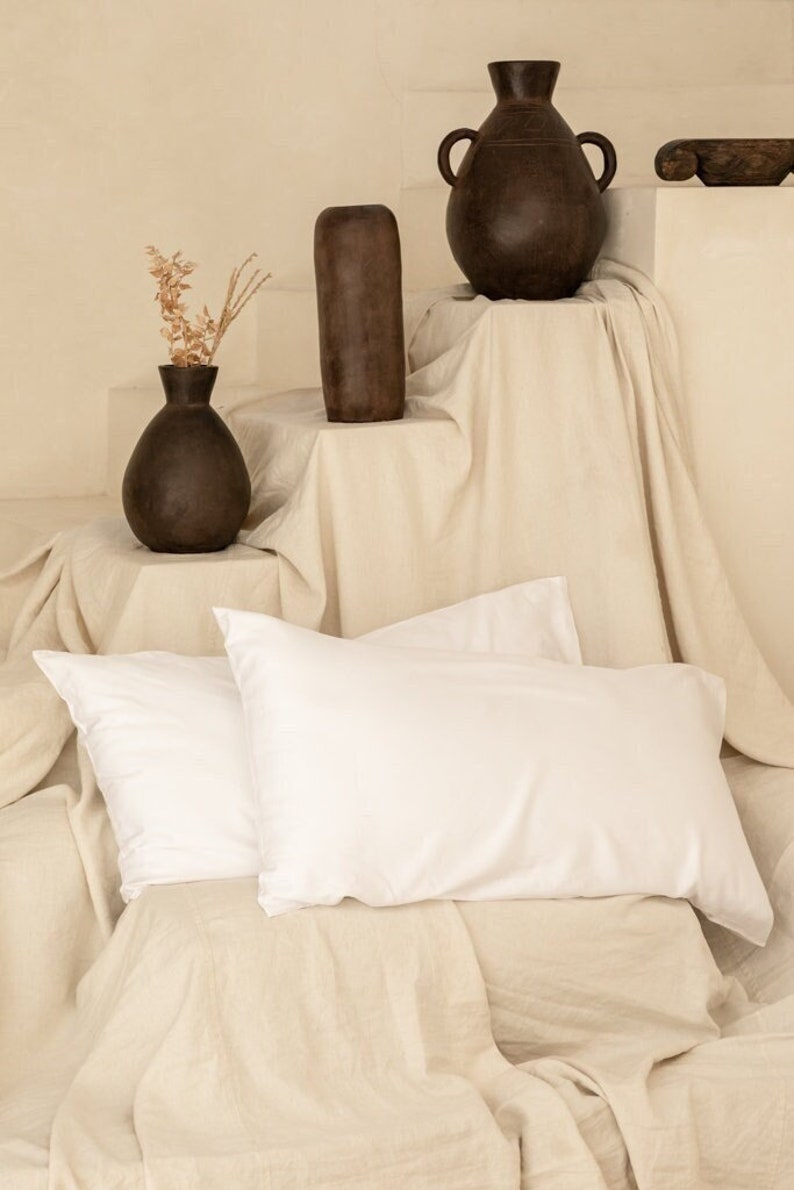 May include: Two white pillowcases on a beige linen fabric background. Three brown ceramic vases are on a shelf behind the pillows.