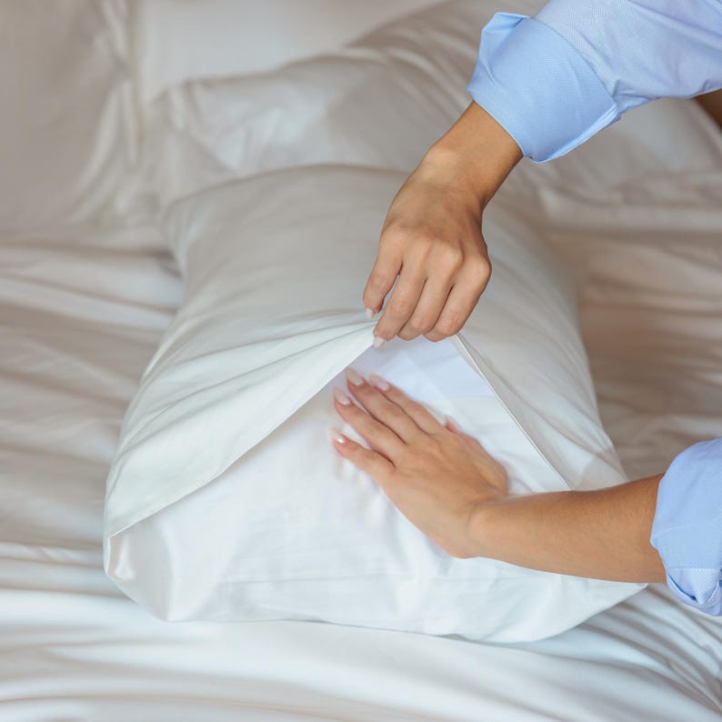 K&ouml;nnte beinhalten: Ein wei&szlig;es Kissen wird in einen wei&szlig;en Kissenbezug gelegt. Das Kissen und der Kissenbezug liegen auf einem wei&szlig;en Bett. Die H&auml;nde der Person sind sichtbar, und sie tr&auml;gt ein hellblaues Hemd.