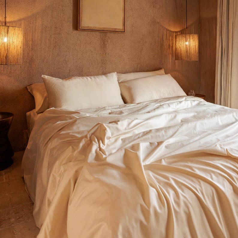 K&ouml;nnte beinhalten: Ein Bett mit cremefarbener Bettw&auml;sche, einschlie&szlig;lich Bettbezug und Kissen. Das Zimmer hat eine strukturierte Wand und zwei H&auml;ngelampen mit gewebten Schirmen. Die Gesamt&auml;sthetik ist warm und einladend.