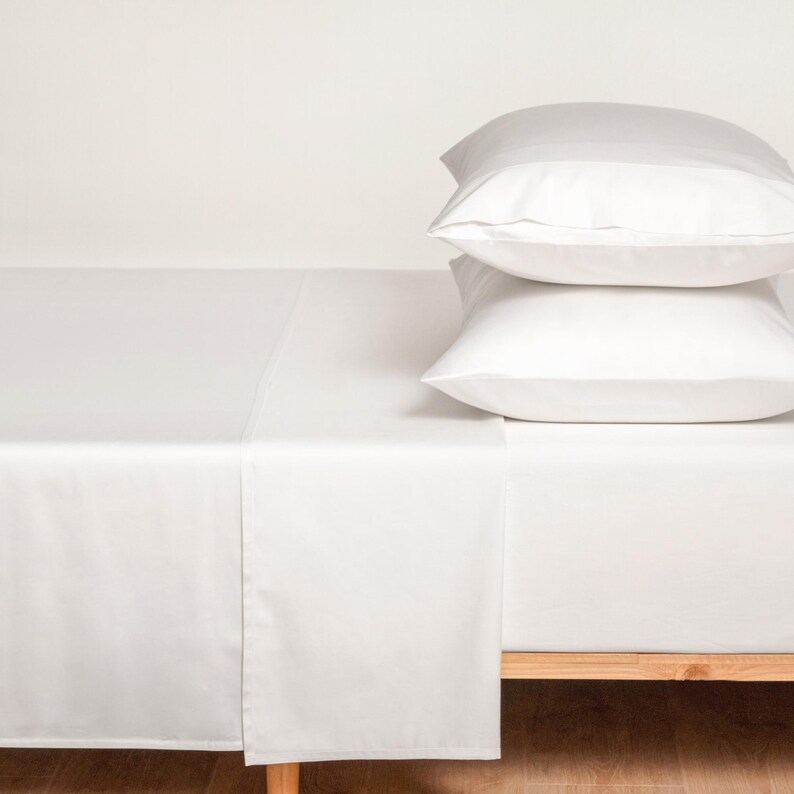 May include: A crisp, white bed sheet set with two matching pillows. The sheets are neatly arranged on a wooden bed frame, creating a clean and minimalist aesthetic. The pillows are stacked on top of the sheets.