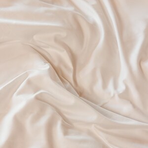 K&ouml;nnte beinhalten: Nahaufnahme eines beigefarbenen Satin-Stoffs mit sanften Falten und dezentem Glanz. Der Stoff wirkt glatt und luxuri&ouml;s, wobei Licht und Schatten Tiefe und Textur erzeugen. Die Gesamt&auml;sthetik ist elegant und minimalistisch.