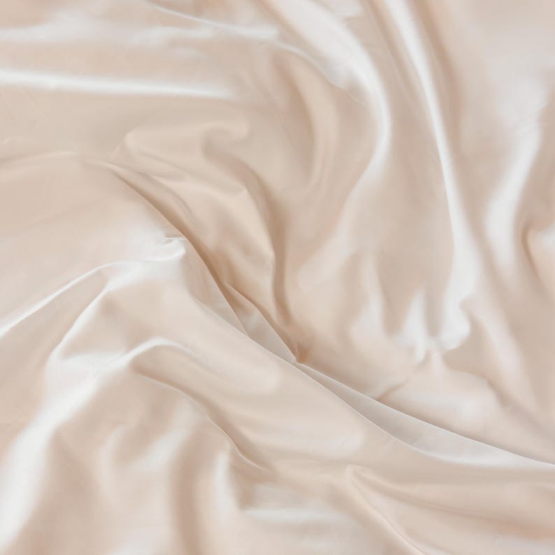 K&ouml;nnte beinhalten: Nahaufnahme eines beigefarbenen Satin-Stoffs mit sanften Falten und dezentem Glanz. Der Stoff wirkt glatt und luxuri&ouml;s, wobei Licht und Schatten Tiefe und Textur erzeugen. Die Gesamt&auml;sthetik ist elegant und minimalistisch.