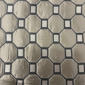 May include: Close-up of a fabric with a repeating geometric pattern. The design features octagons and rectangles in a neutral color palette of gray, white, and dark gray. The fabric has a subtle sheen, suggesting a luxurious material.