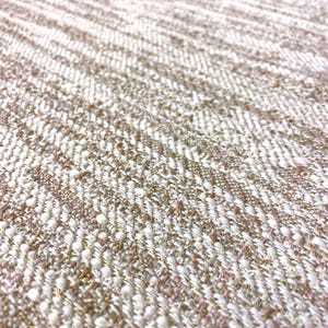 May include: Close-up of a textured fabric with a woven pattern. The fabric features a mix of white, beige, and light pink threads, with subtle gold metallic accents. The weave creates a diagonal striped effect.