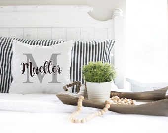 May include: White pillow with a grey letter M and the name "Mueller" in black script. The pillow is on a bed with black and white striped pillows and a wooden tray with a potted plant and wooden beads.