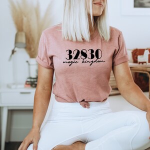 Op de afbeelding: Een vrouw draagt een roze T-shirt met de tekst "32830 magic kingdom" erop gedrukt. Ze zit op een wit tapijt met een witte houten tafel achter haar.