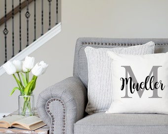 May include: A white throw pillow with a grey letter M and the name "Mueller" in black script. The pillow is on a grey sofa with a striped pillow behind it.