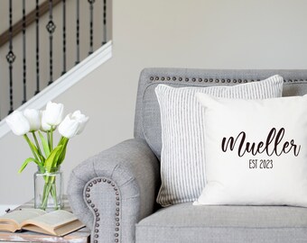 May include: White cushion with black cursive text that reads "Mueller Est 2023" on a grey sofa with a striped cushion.