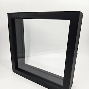 May include: A black shadow box frame with a clear glass front. The frame is square and has a deep profile, creating a three-dimensional effect. The frame is designed to display items.