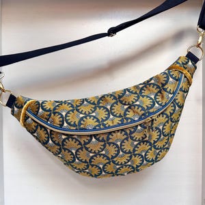 Pode incluir: Uma pochete curva com um padrão repetido de formas de leque douradas e verde-azulado. A bolsa tem um zíper azul, ferragens douradas e uma alça preta ajustável. A bolsa está pendurada em um fundo branco.