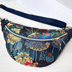 Op de afbeelding: Een marineblauwe heuptas met een bloemenpatroon in goud, rood, blauw en groen. De tas heeft een gouden rits en een marineblauwe verstelbare riem. De tas is gemaakt van een patroonstof.