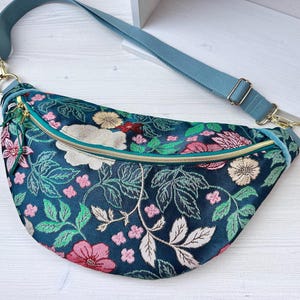 Op de afbeelding: Een donkerblauwe heuptas met een bloemenpatroon met roze, witte en gele bloemen en groene bladeren. De tas heeft een blauwgroene band, goudkleurige hardware en een blauwgroene rits. De tas is gemaakt van een zachte, getextureerde stof.