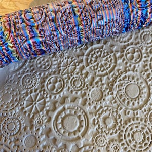 Pode incluir: Um close-up de um rolo de massa com um padrão de engrenagens e círculos. O rolo é feito de madeira e tem um acabamento colorido e iridescente. O rolo está sendo usado para criar uma impressão em uma peça de argila.