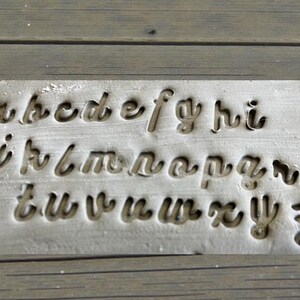 May include: A rectangular metal stamp with the lowercase alphabet in a cursive font. The letters are cut out of the metal, creating a negative space design. The stamp is a light gray color and sits on a wooden surface.