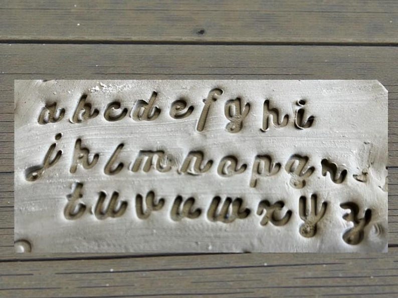 May include: A rectangular metal stamp with the lowercase alphabet in a cursive font. The letters are cut out of the metal, creating a negative space design. The stamp is a light gray color and sits on a wooden surface.