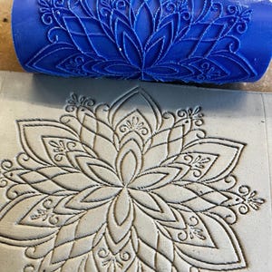 May include: A bright blue cylindrical clay texture roller featuring a raised, intricate mandala pattern. The roller is positioned above a flat, light gray slab of clay that displays a deeply impressed, matching floral mandala design.