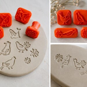 May include: A set of bright orange stamps featuring various bird and flower designs. The stamps have square or rectangular bases with raised patterns on the bottom, shown creating impressions of birds and flowers in a light grey clay surface.