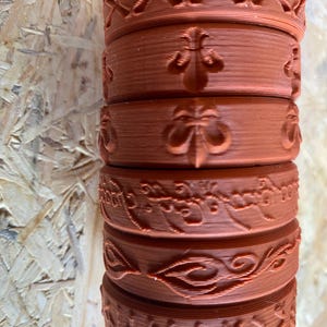 Op de afbeelding: Een stapel van zes bruine 3D-geprinte ringen met verschillende ontwerpen. De bovenste ring heeft een herhalend patroon van X'en. De tweede ring heeft een fleur-de-lis-ontwerp. De derde ring heeft een fleur-de-lis-ontwerp. De vierde ring heeft een herhalend patroon van schrift. De vijfde ring heeft een herhalend patroon van bladeren. De onderste ring heeft een herhalend patroon van X'en.