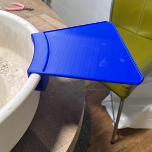 Pode incluir: Uma bandeja de plástico azul brilhante, projetada para ser fixada a um objeto branco e arredondado. A bandeja é montada em uma superfície branca e curva. O fundo inclui uma mesa de madeira, uma cadeira verde e um alicate rosa.