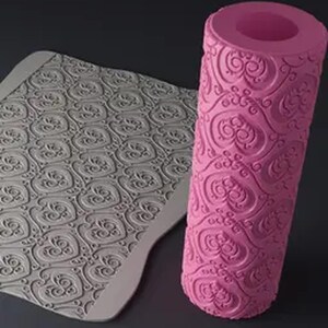 Pode incluir: Um rolo rosa e uma esteira de silicone cinza com um padrão de coração. O rolo é cilíndrico com um design decorativo em relevo. A esteira tem um padrão em forma de coração, ideal para assar. Os itens estão em um fundo escuro.