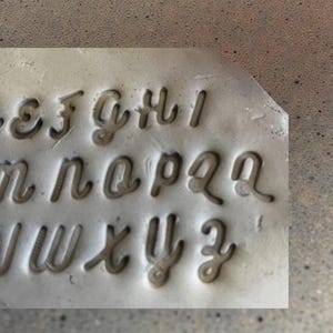 May include: A gray metal stamp with lowercase cursive letters. The letters are deeply carved into the metal, creating a negative space for imprinting. The letters include e, f, g, h, i, n, o, p, q, r, v, w, x, y, and z.