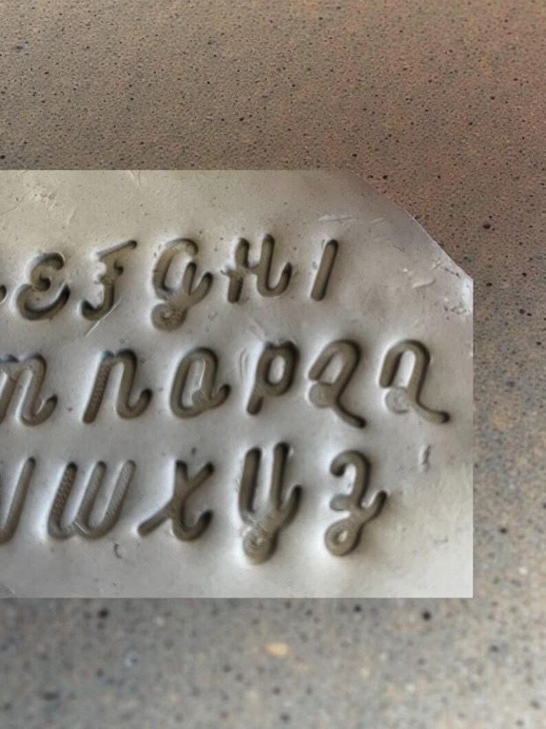 May include: A gray metal stamp with lowercase cursive letters. The letters are deeply carved into the metal, creating a negative space for imprinting. The letters include e, f, g, h, i, n, o, p, q, r, v, w, x, y, and z.