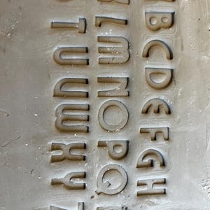 Könnte beinhalten: Eine hellgraue Beton- oder Tonplatte mit dem englischen Alphabet in drei vertikalen Spalten. Die Buchstaben A bis Z sind deutlich in die Oberfläche eingeprägt und zeigen eine glatte, matte Textur mit subtilen Schatten in jedem Zeichen.