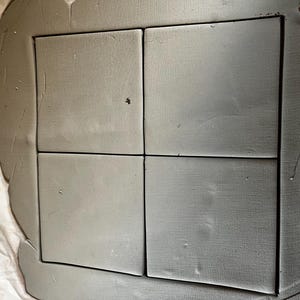 Può includere: Quattro piastrelle quadrate grigie inserite in un bordo grigio. Le piastrelle sono disposte in una griglia 2x2. Le piastrelle e il bordo hanno un aspetto strutturato, che suggerisce un materiale fatto a mano o naturale. Lo sfondo è un tessuto bianco.