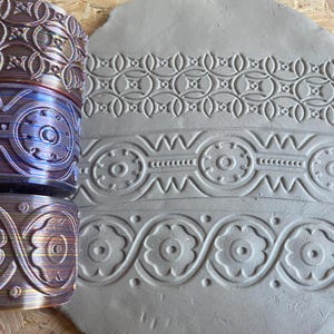Könnte beinhalten: Drei verschiedene gemusterte Nudelhölzer für Ton, jedes mit einem anderen Design. Die Nudelhölzer sind aus Metall und haben eine glänzende, schillernde Oberfläche. Die Designs sind kompliziert und detailliert und wären perfekt, um jedem Tonprojekt eine dekorative Note zu verleihen.