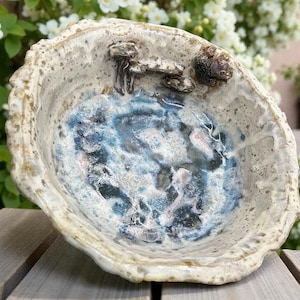 Puede incluir: Un cuenco de cerámica hecho a mano con una forma orgánica. El cuenco presenta un exterior texturizado de color blanquecino y un interior azul y blanco. Pequeñas decoraciones de setas están adheridas al borde. El cuenco tiene unos 15 cm de diámetro.