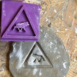 May include: A purple plastic triangle-shaped stamp with a wolf silhouette inside. The stamp is being used to create an impression of the wolf in a gray clay circle.