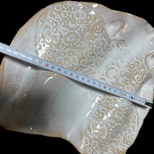 Pode incluir: Uma travessa de cerâmica branca suja, feita à mão, com um padrão semelhante a renda. A travessa tem aproximadamente 28 cm de largura. A borda tem um design ondulado. O fundo é preto.