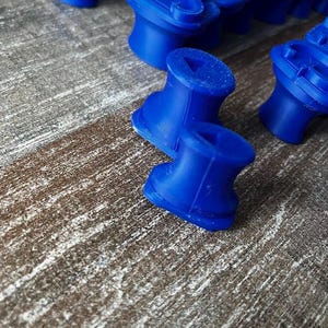May include: Close-up of several bright blue, 3D-printed objects. The objects have a unique shape, with a cylindrical base and a wider top section. The objects are arranged on a textured, brown surface.