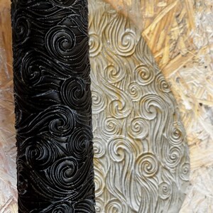 May include: A black rolling pin with a swirling wave pattern design. The rolling pin is used to create a decorative pattern on a surface, such as clay or dough.