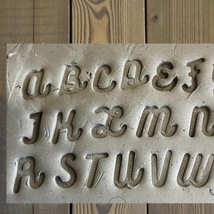 May include: A rectangular, gray stone block with the letters of the alphabet carved into it. The letters are in a cursive font and are deeply etched into the stone. The block is set against a wooden background.