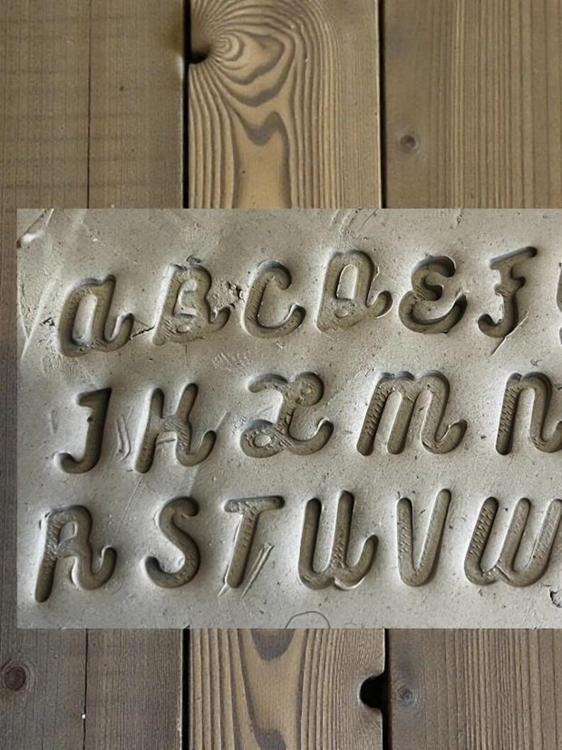 May include: A rectangular, gray stone block with the letters of the alphabet carved into it. The letters are in a cursive font and are deeply etched into the stone. The block is set against a wooden background.