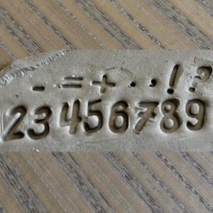 May include: A gray, rectangular stamp with the numbers 1 through 0 and mathematical symbols including minus, equals, plus, period, comma, exclamation point, and question mark. The numbers and symbols are indented.