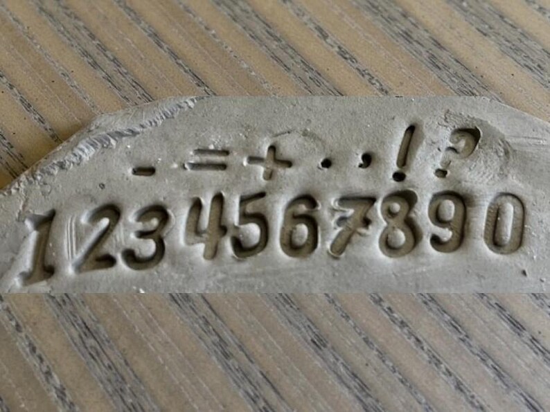 May include: A gray, rectangular stamp with the numbers 1 through 0 and mathematical symbols including minus, equals, plus, period, comma, exclamation point, and question mark. The numbers and symbols are indented.