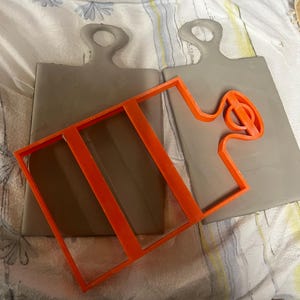 Op de afbeelding: Twee grijze klei snijplanken met gaten voor handgrepen. Een oranje rechthoekig frame en een gebogen oranje vorm zijn op de planken geplaatst. De planken liggen op een wit en geel patroon doek.