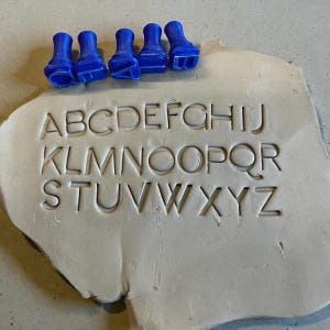 Op de afbeelding: Een stuk lichtgekleurde klei met het alfabet erin gedrukt. Vijf blauwe letterstempels zijn boven de klei geplaatst. De letters A tot en met Z zijn zichtbaar. De klei is ongeveer 15 cm breed.
