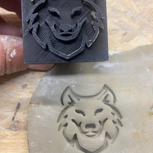 May include: A black 3D printed wolf head stamp is used to create an impression of the wolf head in clay.