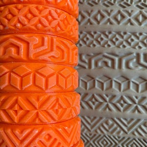 Op de afbeelding: Een close-up van een oranje deegroller met meerdere verwisselbare ontwerpen. De deegroller is gemaakt van plastic en heeft een glad oppervlak. De ontwerpen zijn geometrisch en omvatten een verscheidenheid aan patronen.