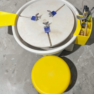 Könnte beinhalten: Eine Töpferscheibe mit einer weißen, kreisförmigen Arbeitsfläche, blauen Klemmen und einem gelben Werkzeughalter. Darunter befindet sich ein gelber Deckel. Das Rad ist für Keramik und Töpferei.
