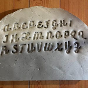 May include: A gray, semi-circular slab with the alphabet carved into it. The letters are in a stylized, cursive font. The slab is set against a wooden background.