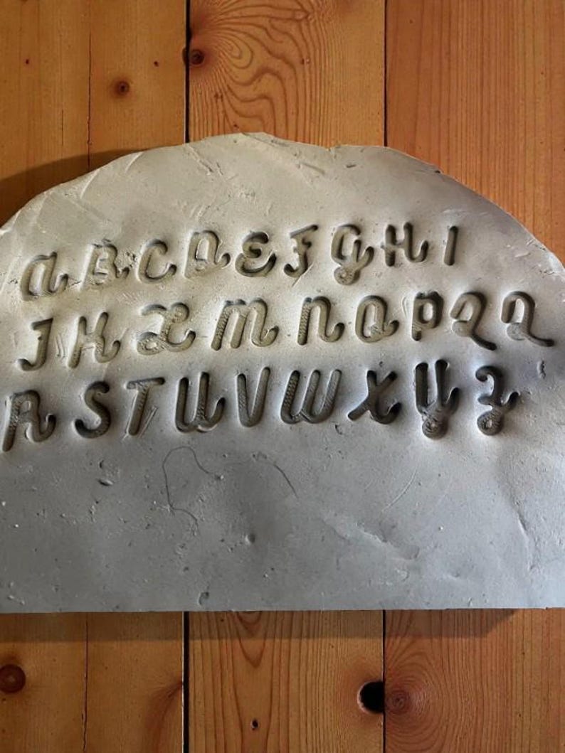 May include: A gray, semi-circular slab with the alphabet carved into it. The letters are in a stylized, cursive font. The slab is set against a wooden background.