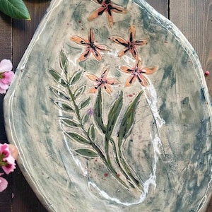 Puede incluir: Una fuente de cerámica hecha a mano, de forma ovalada. La fuente es principalmente de color verde azulado apagado con un acabado moteado. Presenta un diseño floral pintado a mano con flores naranjas y hojas verdes.