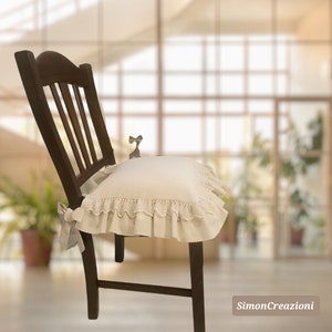 Op de afbeelding: Een wit stoelkussen met een kantversiering en een strik aan de achterkant. Het kussen ligt op een houten stoel voor een wazige achtergrond.